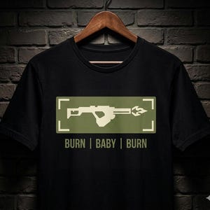 Könnte beinhalten: Schwarzes T-Shirt mit einem grünen Rechteck, das eine weiße Waffengrafik und den Text "BURN | BABY | BURN" enthält. Das Design zeigt eine Waffe, die eine Flamme abfeuert. Das T-Shirt hängt an einem Holzkleiderbügel.