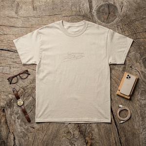 May include: A beige t-shirt with the text "FIND ME IN THE WAVES" in a minimalist design. The shirt is displayed on a wooden surface, alongside a pair of glasses, a watch, a phone, and a charging cable.