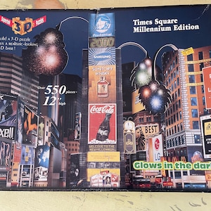 Op de afbeelding: Een doos met 3D-puzzel van Times Square, Millennium Edition, met meer dan 550 stukjes. De doos toont een nachtscène met verlichte billboards, waaronder Coca-Cola en Prudential 2000. De puzzel gloeit in het donker en is 30 cm hoog.