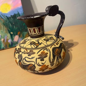 May include: A ceramic pitcher featuring a cream-colored base with black and orange hand-painted motifs. The design includes stylized animals, floral patterns, and geometric bands around the neck. It has a single handle and a wide, flared rim.