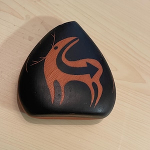 May include: A triangular-shaped stone with a smooth black surface featuring a stylized terracotta-colored deer illustration. The deer design includes a prominent arrow shape integrated into its body, with thin lines depicting antlers on the left side.
