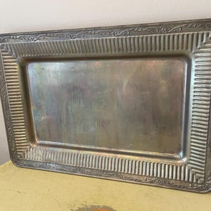 May include: A rectangular silver-toned metal serving tray featuring a smooth, flat center surface surrounded by a wide, textured border. The border displays a vertical ribbed pattern and an outer edge decorated with a delicate, embossed floral vine motif.