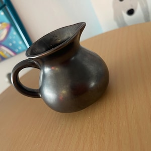 May include: A small, rounded ceramic pitcher with a smooth, polished black finish. The vessel features a curved handle on the side and a distinct pouring spout at the top rim, showcasing a uniform dark metallic sheen across its entire surface.