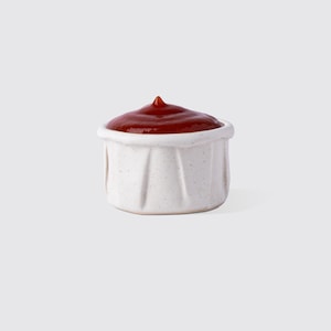 Peut inclure: Un petit bol à condiments en céramique blanche rempli de ketchup rouge. Le bol a une surface texturée et mouchetée et un design unique et plié. Le ketchup est d'un rouge profond, avec un petit pic au sommet.