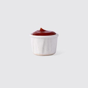 Op de afbeelding: Een kleine, witte keramische condimentbeker gevuld met rode ketchup. De beker heeft een gespikkelde textuur en een enigszins onregelmatige vorm. De ketchup is glad en glanzend, met een kleine piek aan de bovenkant.