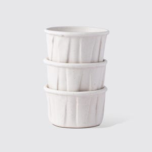 May include: Three stacked, off-white ceramic ramekins. Each ramekin has a ridged exterior and a rounded rim. The speckled finish adds texture to the simple, yet elegant design. These small bowls are ideal for baking or serving individual portions.