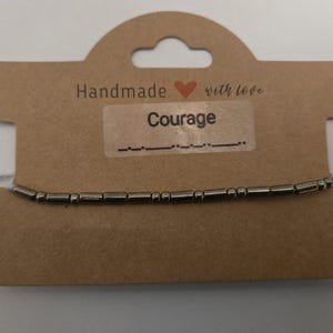 Könnte beinhalten: Ein handgefertigtes Armband mit dem Wort "Courage" in Morsecode. Das Armband besteht aus kleinen, silberfarbenen Perlen, die auf eine dünne Schnur aufgefädelt sind. Es wird auf einer braunen Karte mit der Aufschrift "Handmade with love" präsentiert.