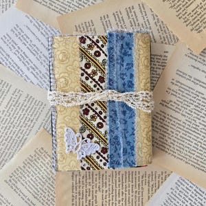 May include: A rectangular journal cover featuring vertical fabric strips in cream with floral patterns, brown and cream geometric prints, and blue with floral motifs. A white crocheted lace butterfly is attached, and a cream lace ribbon wraps around.