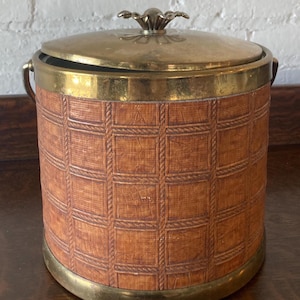 Op de afbeelding: Vintage ijsemmer met een messing deksel en afwerking. De cilindrische behuizing is bedekt met een getextureerd, bruin materiaal met een rasterpatroon. Het heeft een decoratieve messing handgreep en dekselknop. Een stijlvolle toevoeging aan elke bar.