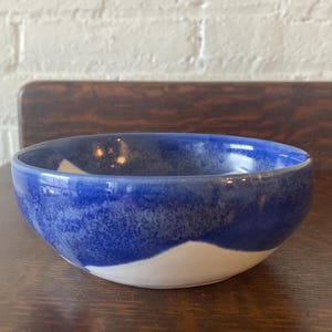Puede incluir: Un cuenco de cerámica hecho a mano con un acabado brillante. El cuenco presenta un diseño llamativo con una sección superior azul oscuro y una sección inferior blanca. Ideal para servir aperitivos o comidas pequeñas.