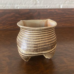 May include: A small, handcrafted ceramic pot with a square top and rounded body. The pot is tan with horizontal brown stripes and rests on three small feet. The interior is a lighter tan color. The pot is likely used for plants or small items.