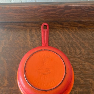 Könnte beinhalten: Kleine, runde, rot-orange emaillierte Gusseisenpfanne mit Griff. Die Pfanne hat einen leuchtend orangefarbenen Innenraum und einen Farbverlauf von Rot und Orange auf der Außenseite. Auf dem Boden steht "Made in Belgium".