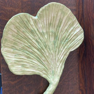 Puede incluir: Un plato de cerámica verde claro con forma de hoja y vetas texturizadas. El plato tiene una forma curvada y una base en forma de tallo. El color es un verde suave y apagado con sutiles variaciones de tono. El plato probablemente se utiliza con fines decorativos o de servicio.