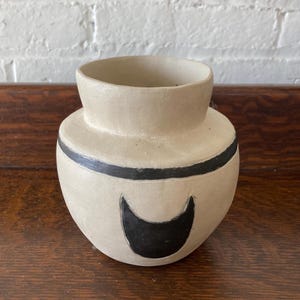 May include: A small, round, off-white ceramic vase with a wide opening and a short neck. The vase features a horizontal black line encircling the upper body and a prominent black crescent moon shape painted on the front center of the rounded base.
