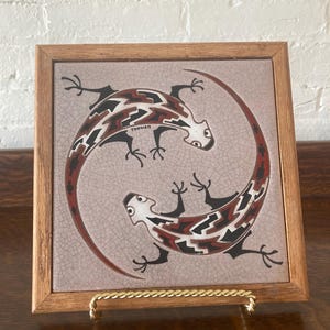 May include: Square framed tile featuring two stylized lizards in a circular design. The lizards are black, white, and red, set against a light gray background. The frame is made of wood. The tile is displayed on a gold-colored metal stand.