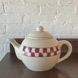 Puede incluir: Tetera de cerámica color crema con tapa y asa. La tetera presenta una banda horizontal de cuadrados rojos y blancos alternados alrededor de su cuerpo. El pico y el asa son curvos.