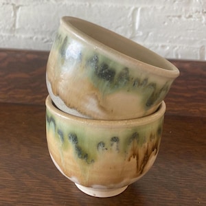 May include: Two handmade ceramic bowls stacked on top of each other. The bowls have a unique glaze with a blend of green, tan, and brown colors. The bowls are sitting on a dark wooden surface.