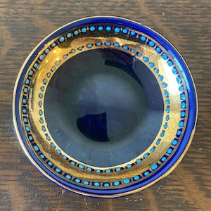 May include: A small, round, blue dish with a gold rim and accents. The rim features a repeating pattern of small, turquoise-colored dots. The interior of the dish is a deep, glossy blue.