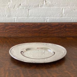 May include: A small, silver-colored plate with a decorative rim and a curved design on the interior. The plate is oval-shaped and reflects the light, suggesting a polished surface. The plate is likely made of metal and is suitable for serving small portions.
