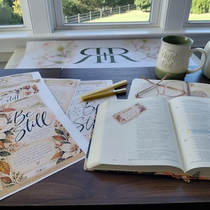 May include: An open Bible with a floral bookmark, two yellow pens, a white ceramic mug with green trim, a white pumpkin, and several printed devotional sheets featuring the text Be Still and floral illustrations on a dark wooden surface.