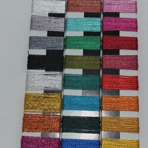 May include: A vertical arrangement of colorful metallic thread spools. The spools are in various colors, including silver, gold, blue, green, and pink. Each spool is neatly wound and presented on a white card with a black top.