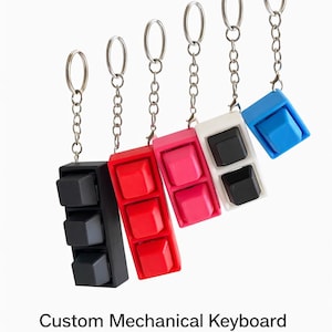 Könnte beinhalten: Schlüsselanhänger in Form von mechanischen Tastaturtasten in Schwarz, Rot, Pink, Weiß und Blau. Jeder Schlüsselanhänger hat eine silberne Kette und einen Schlüsselring. Der Text "Custom Mechanical Keyboard Keychain" steht darunter.