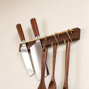 May include: A wooden kitchen utensil rack with brass hooks and a brass bar. Two stainless steel knives with brown leather handles are mounted on the rack. Three wooden spoons hang from the hooks. The rack is mounted on a cream wall.