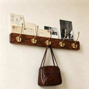 May include: A wooden wall-mounted coat rack with five brass hooks. The rack has a dark brown finish and a small shelf holding vintage postcards and a brown leather handbag hanging from one of the hooks.
