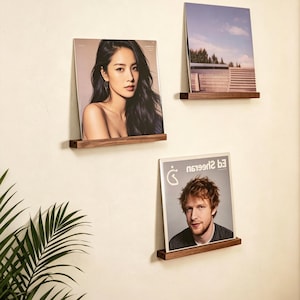May include: Three dark brown wooden floating shelves mounted on a wall, each holding a square album cover. The top left shelf displays a portrait of a woman, the top right shelf shows a landscape, and the bottom right shelf features an Ed Sheeran album.
