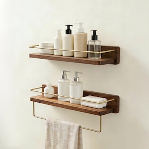 May include: Two wall-mounted wooden shelves with gold-toned metal railings. The top shelf holds four assorted bottles and a jar. The bottom shelf holds two pump bottles, a white rectangular soap bar, and a towel hanging from a gold metal bar.