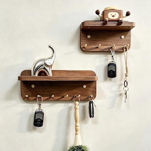 May include: Two wooden key holder shelves with brass hooks. One shelf has a silver elephant figurine, the other has a wooden cartoon figure. Keys and a brush hang from the hooks. The shelves are mounted on a white wall.