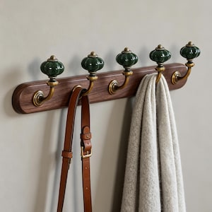 May include: A wall-mounted coat rack featuring a horizontal dark wood base with five brass hooks. Each hook is topped with a round, ribbed, dark green ceramic knob. A brown leather bag strap and a light gray knit scarf hang from the hooks.