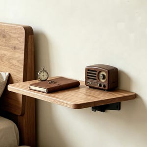 May include: A wooden floating shelf with a brown leather-bound journal, a vintage pocket watch, and a small, brown retro radio. The shelf is mounted on a white wall next to a wooden bed frame.