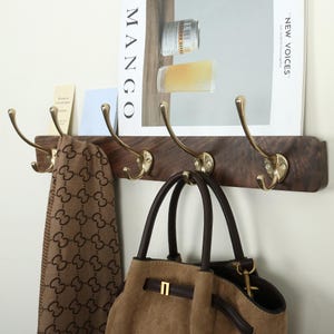 May include: A wall-mounted coat rack featuring a dark brown wooden board with four polished gold-tone metal hooks. A brown patterned scarf hangs from the leftmost hook, and a tan suede handbag with dark brown handles hangs from the center.