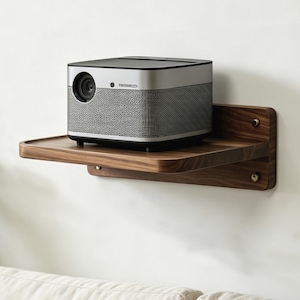 Op de afbeelding: Een zilveren en grijze projector met een zwart bovenstuk staat op een donkere houten plank. De plank is aan een witte muur bevestigd. De projector heeft een lens en een getextureerde grijze zijkant.