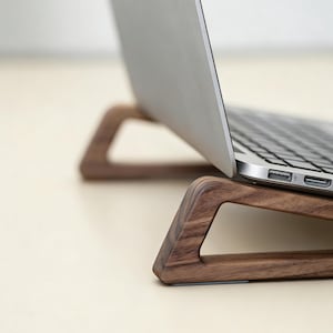 Può includere: Un supporto per laptop in legno con finitura a venatura naturale. Il supporto ha un design triangolare con un ritaglio per il flusso d'aria e supporta un laptop argentato. Il supporto è progettato per sollevare il laptop.