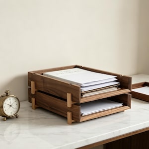 Pode incluir: Um organizador de mesa de madeira de dois níveis com acabamento natural. O organizador contém pilhas de papel e documentos brancos. O design apresenta uma estética minimalista, adequada para uso em escritório ou em casa. Um pequeno relógio analógico dourado está à esquerda.