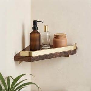 May include: A triangular corner shelf made of dark brown wood with a brushed gold metal railing. On the shelf sits a brown glass soap dispenser with a black pump, a clear glass bottle with a gold cap, and a small matte brown jar with a cork lid.