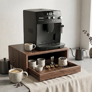 May include: A black De'Longhi Magnifica espresso machine sits atop a dark wood storage unit with a pull-out drawer. The drawer contains small ceramic cups, a metal tamper, gold spoons, and a fabric bag of coffee beans. Two mugs sit nearby.