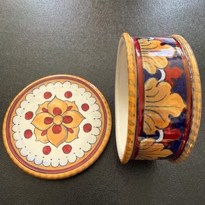 Op de afbeelding: Een keramische container met een deksel, met een bloemenontwerp in geel, rood en blauw. Het deksel is rond met een geschulpte rand en een centraal bloemmotief. De container heeft een cilindrisch lichaam met vergelijkbare bloempatronen.