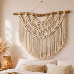 Puede incluir: Un gran tapiz de macramé en color blanco roto suspendido de una rama de madera natural. El diseño presenta varias filas superpuestas en semicírculo de cuerda anudada y cuentas de madera redondas, con largos flecos rectos que cuelgan.
