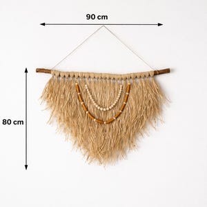 K&ouml;nnte beinhalten: Ein Wandbehang aus Naturfasern, 90 cm breit und 80 cm lang, mit einem Holzd&uuml;bel und dekorativen Holzperlen. Der beigefarbene Fransenbehang h&auml;ngt in einer dreieckigen Form und erzeugt eine Boho-&Auml;sthetik.