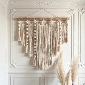 Pode incluir: Uma grande tapeçaria de parede em macramé creme. A peça é suspensa por uma viga de madeira natural e apresenta longas mechas verticais de algodão atado. Contas e borlas de madeira branca adicionam textura e detalhes à decoração em estilo boêmio.
