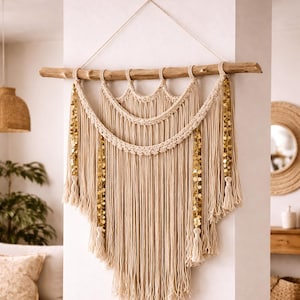 May include: A beige macrame wall hanging with a wooden dowel. The design features layered, fringed sections with gold sequin accents and tassels. This bohemian-style decor adds texture and warmth to any space.