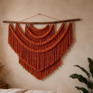 Könnte beinhalten: Ein Wandbehang aus Makramee in gebranntem Orange, der an einem horizontalen Holzast hängt. Das Stück besteht aus drei geschichteten, halbkreisförmigen Abschnitten aus geknoteter Kordel, die in langen, fließenden Fransen in V-Form enden.