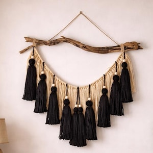 May include: A handcrafted wall hanging featuring a natural wood branch, beige macrame, and black tassels. The decorative piece includes wooden beads and a neutral color palette, suitable for bohemian or modern decor.