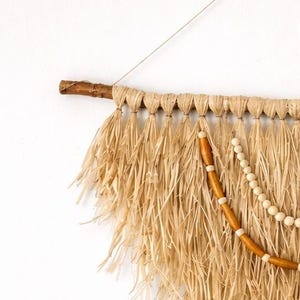 K&ouml;nnte beinhalten: Wandbehang aus Naturfasern mit rustikaler &Auml;sthetik. Er verf&uuml;gt &uuml;ber eine Basis aus einem Holzast, gewebte Raffia-Fransen und dekorative Holzperlen. Die neutralen T&ouml;ne und Texturen erzeugen einen Bohemian-Vibe, passend f&uuml;r die Inneneinrichtung.