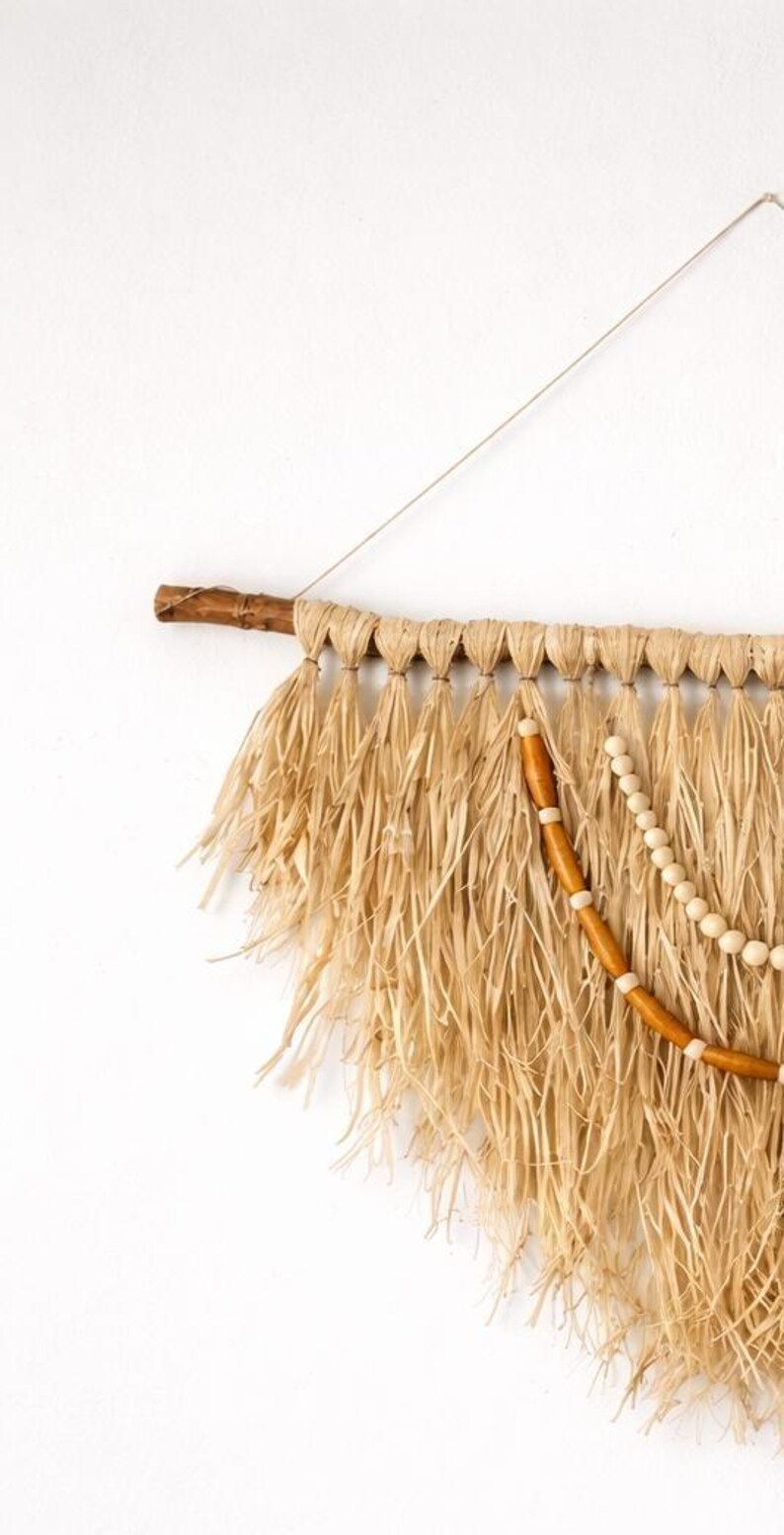 K&ouml;nnte beinhalten: Wandbehang aus Naturfasern mit rustikaler &Auml;sthetik. Er verf&uuml;gt &uuml;ber eine Basis aus einem Holzast, gewebte Raffia-Fransen und dekorative Holzperlen. Die neutralen T&ouml;ne und Texturen erzeugen einen Bohemian-Vibe, passend f&uuml;r die Inneneinrichtung.