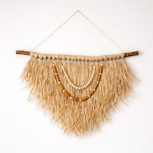 K&ouml;nnte beinhalten: Ein Wandbehang aus Naturfasern mit dreieckiger Form. Die Wanddekoration hat einen braunen Holzast oben, mit einem gewebten Strohdesign und zwei Perlenstr&auml;ngen in der Mitte. Die Perlen sind hellbraun und wei&szlig;.
