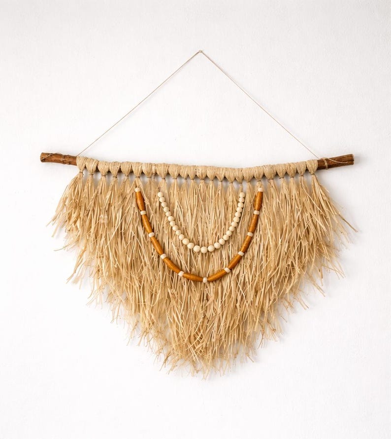 K&ouml;nnte beinhalten: Ein Wandbehang aus Naturfasern mit dreieckiger Form. Die Wanddekoration hat einen braunen Holzast oben, mit einem gewebten Strohdesign und zwei Perlenstr&auml;ngen in der Mitte. Die Perlen sind hellbraun und wei&szlig;.